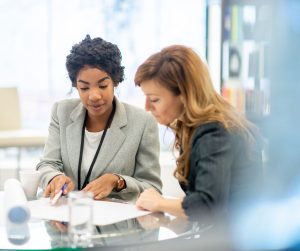 Two women discussing work permit eligibility requirements in Canada, one pointing to documents on a table.